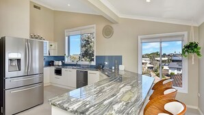 A modern kitchen with stainless steel appliances, a marble countertop, and large windows offering a view of the neighborhood. - The Sea Serenade - Coastal haven with panoramic ocean views! (Swansea Heads)