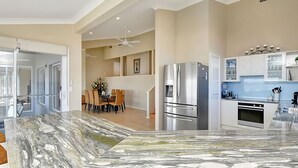 A modern kitchen with a marble countertop, stainless steel appliances, and a dining area with chairs and a table. - The Sea Serenade - Coastal haven with panoramic ocean views! (Swansea Heads)
