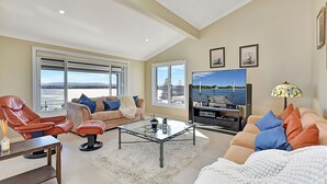 A spacious living room with a large window, a flat-screen TV, and a cozy seating area. - The Sea Serenade - Coastal haven with panoramic ocean views! (Swansea Heads)