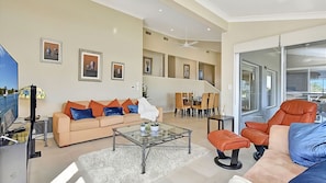 Un salon comprenant un canapé, une table basse et des fauteuils, un coin repas avec une table et des chaises, et une grande fenêtre.