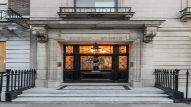 Entrada de un edificio con una puerta negra, un banco pequeño y una lámpara sobre la puerta.