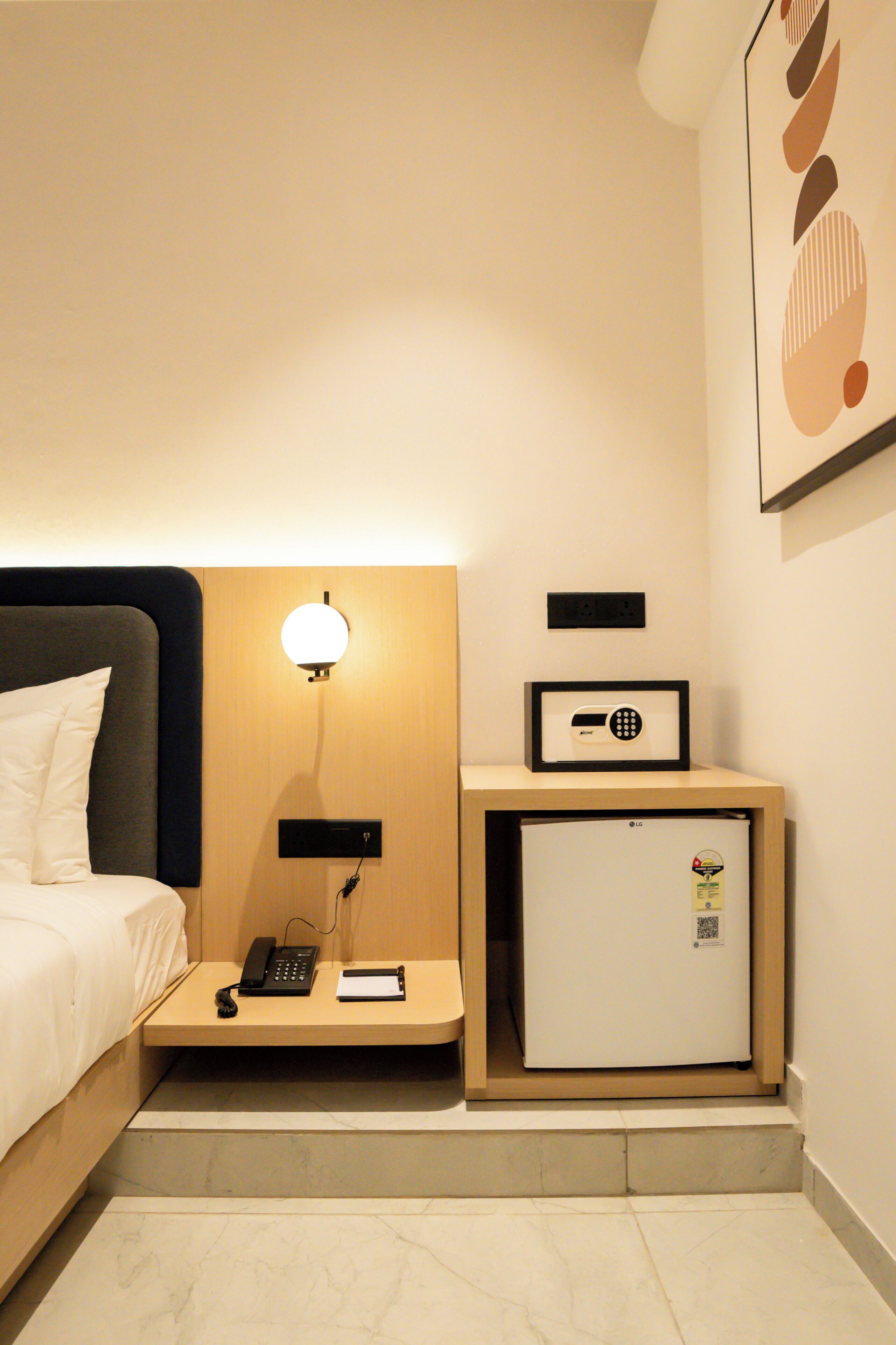 Habitación de hotel con cama, mesita de noche con teléfono, lámpara y un refrigerador pequeño.