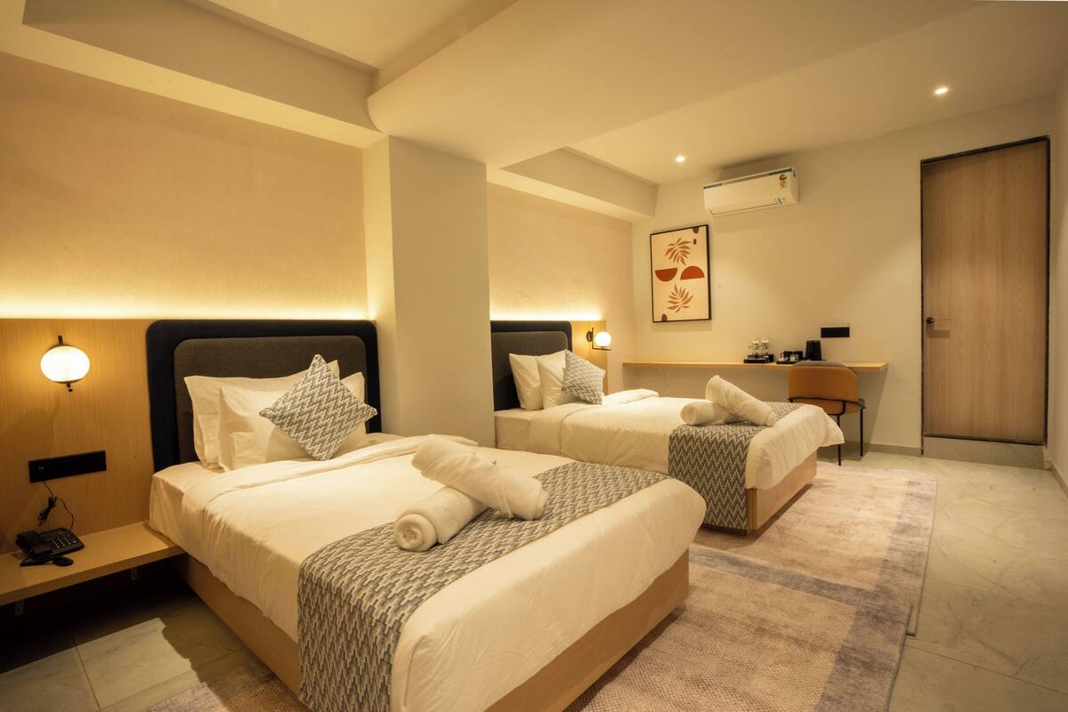 A hotel room with two beds, a desk, and a chair. There is a phone on the nightstand and an air conditioning unit on the wall.