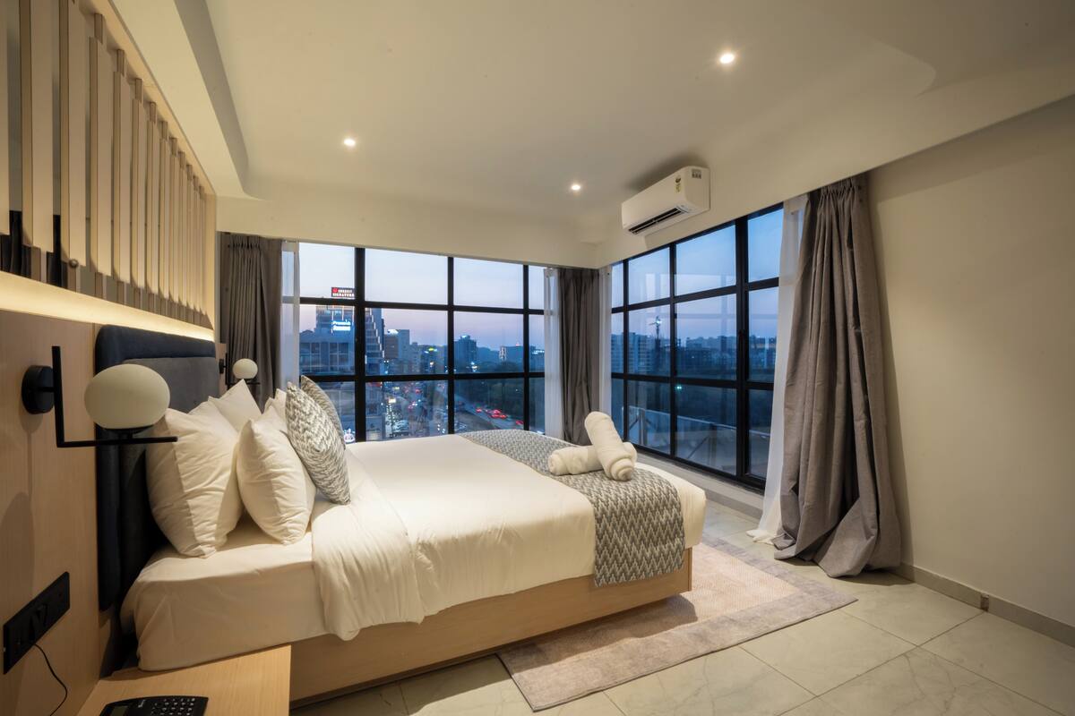 A modern bedroom with a large bed, city view, and an air conditioning unit.