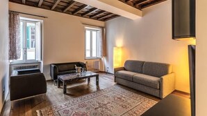 A living room with a sofa, a coffee table, and a patterned rug. There are windows with curtains, a radiator, and a wooden ceiling. - Domus Mondrian - Apartment in Central Roma (Roma)