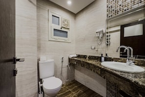 Et moderne badeværelse med toilet, vask og brusebad. Der er et vindue med en aircondition over toilettet, et håndklæde hænger på væggen og et spejl over vasken.