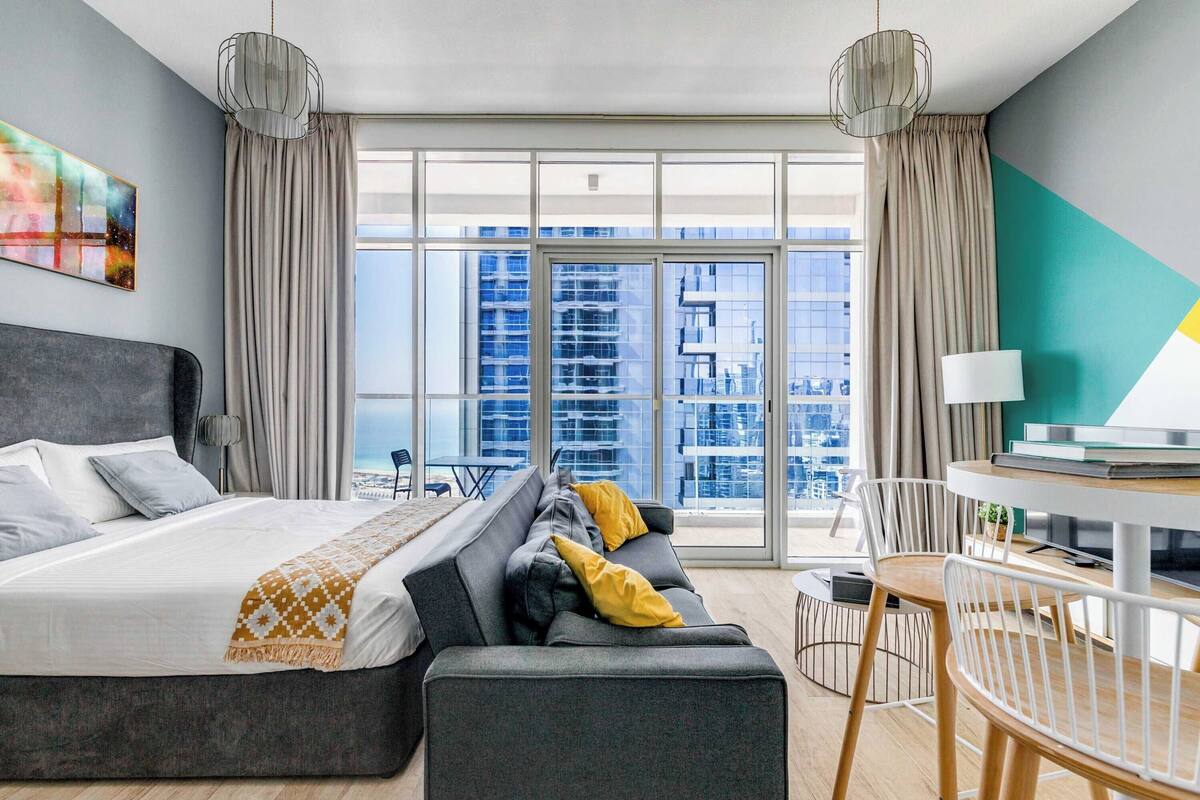 A modern hotel room with a large bed, a sofa, a dining area, and a view of the cityscape through large windows.