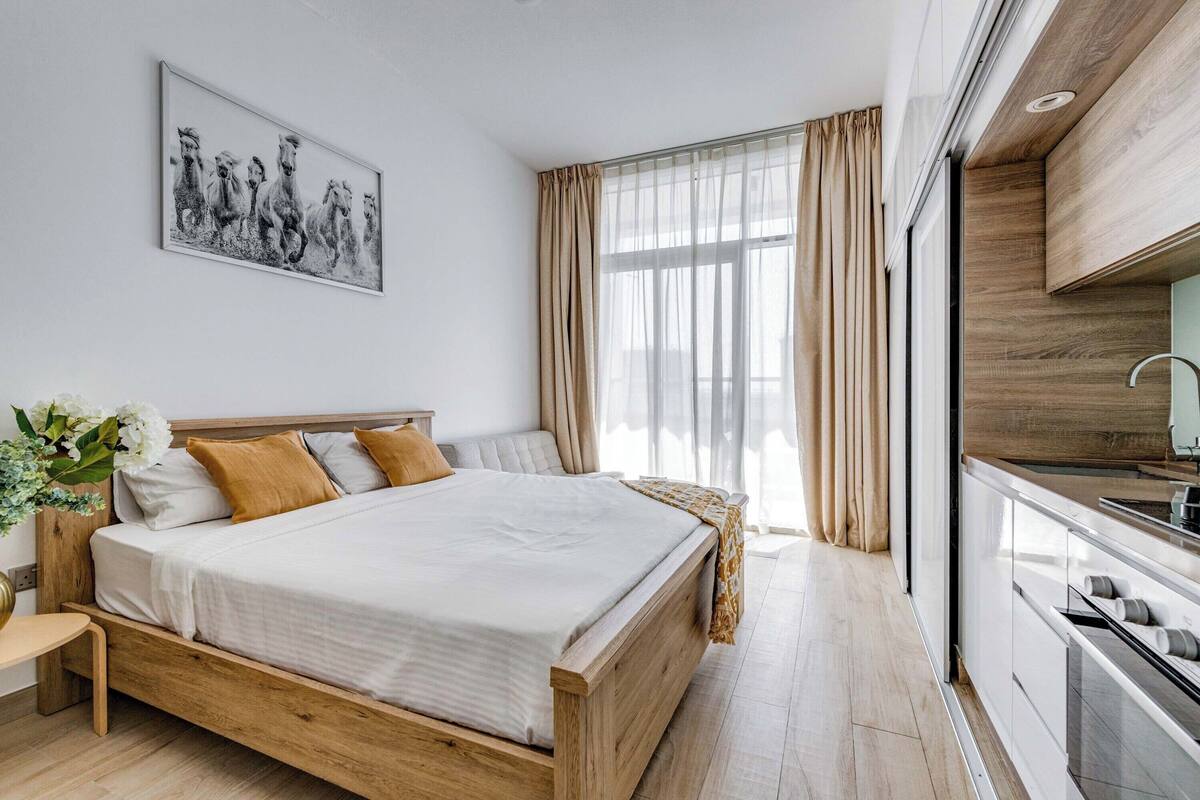 A modern bedroom with a wooden bed, a large window with curtains, and a kitchen area with a sink and stove.