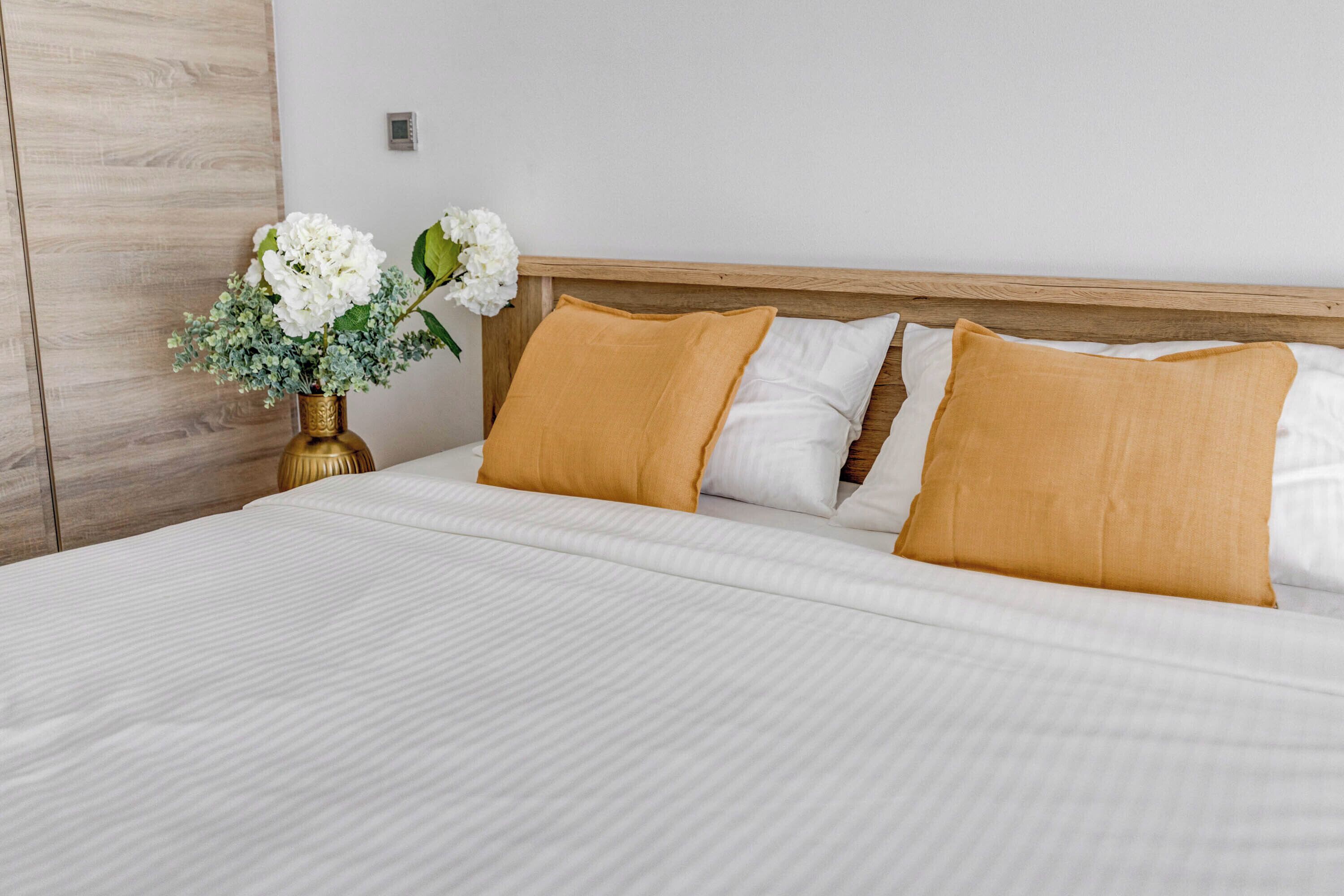 Uma cama bem arrumada com roupa de cama branca e dois travesseiros amarelo-mostarda, um vaso com flores brancas e uma cabeceira de madeira.