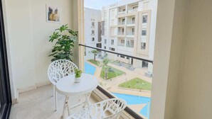 A balcony with white chairs and a small table, overlooking a swimming pool and adjacent buildings.