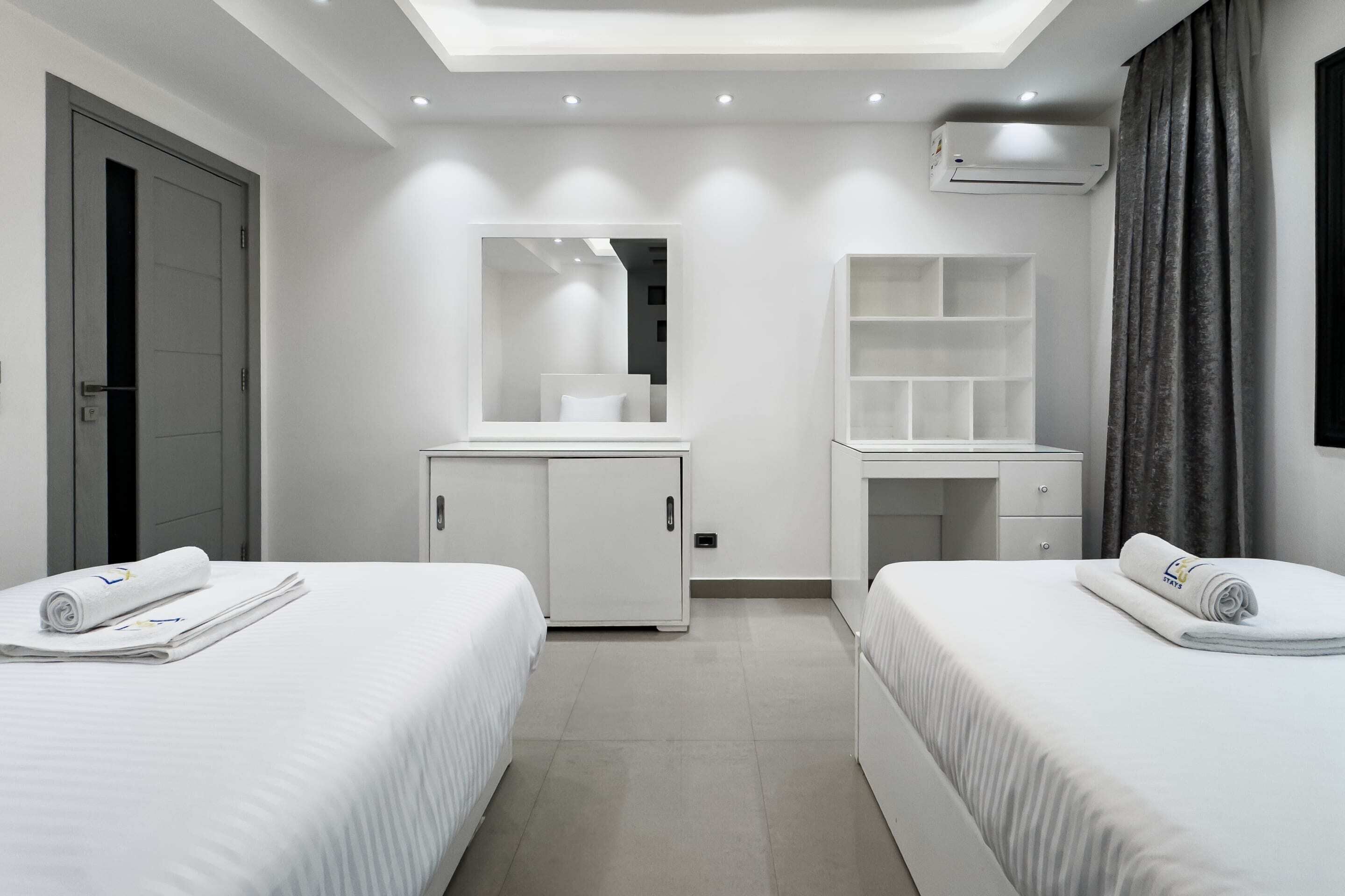 Ein modernes Hotelzimmer mit zwei Betten, einem zentralen Kleiderschrank, einem Schminktisch mit Spiegel und einer eingebauten Regaleinheit.