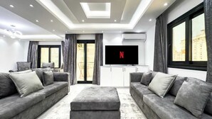 A modern living room with grey sofas, a central ottoman, and a flat-screen TV on a white cabinet. - Xuru Stays I The Bold & Bright 3bdr Apt. 15 Min To Airport (Cairo)
