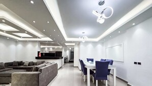 A modern living room with a sectional sofa, a dining table with chairs, recessed ceiling lighting, and a kitchen area in the background. - Xuru Stays I The Bold & Bright 3bdr Apt. 15 Min To Airport (Cairo)