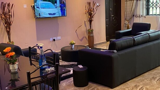 A modern living room with a black sofa, a TV mounted on the wall, a dining table with chairs, and a decorative vase. - Ekoara Apartment (Sekondi-Takoradi)
