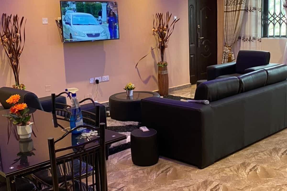 A modern living room with a black sofa, a TV mounted on the wall, a dining table with chairs, and a decorative vase.