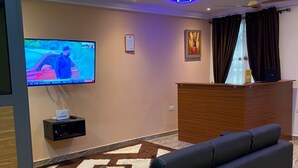 A modern living room with a black sofa, a flat-screen TV mounted on the wall, a wooden reception desk, and a patterned rug. - Ekoara Apartment (Sekondi-Takoradi)