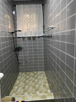 Un petit espace de douche carrelé, équipé d’un pommeau de douche, d’un rideau et d’une petite étagère sur laquelle se trouve une bouteille verte.