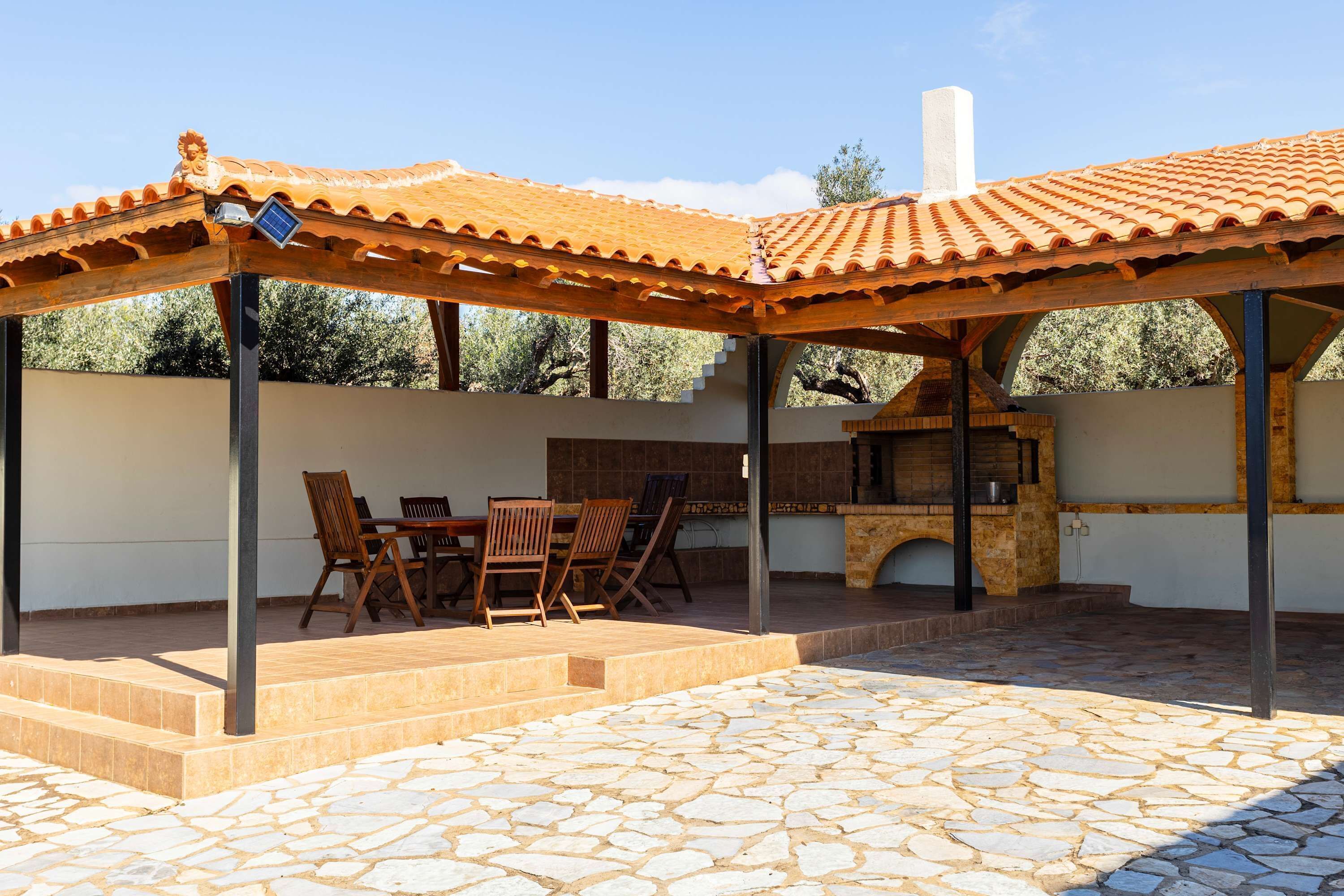 Un patio exterior con suelo de piedra, mobiliario de madera y estructura de techo con tejas.