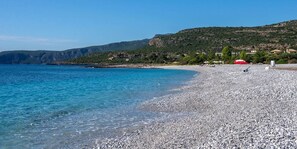 A pebble beach with clear blue water, a red umbrella, and a hillside in the background.