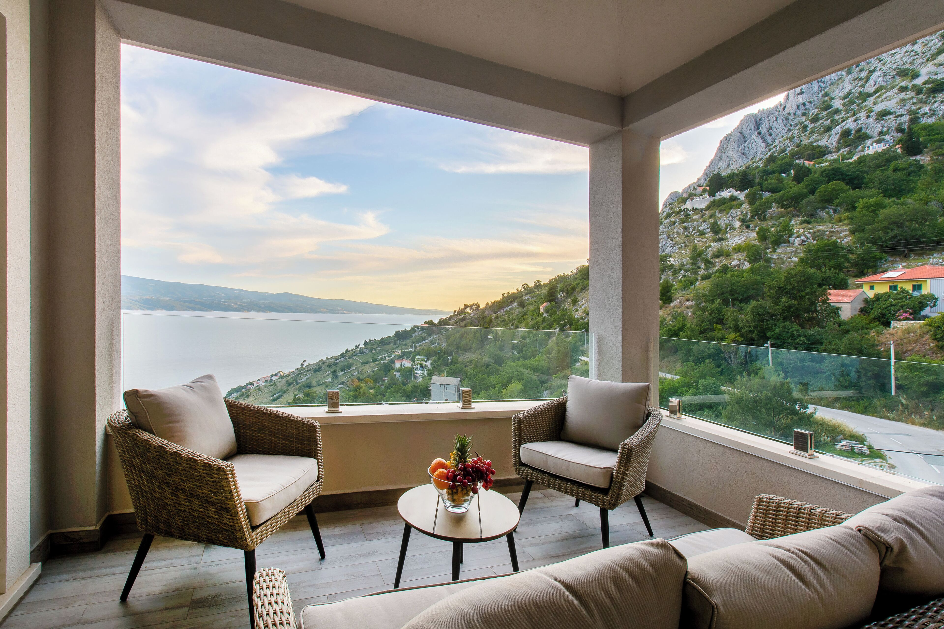 A balcony with wicker furniture, a small table with a fruit bowl, and a view of a mountainous landscape and water.