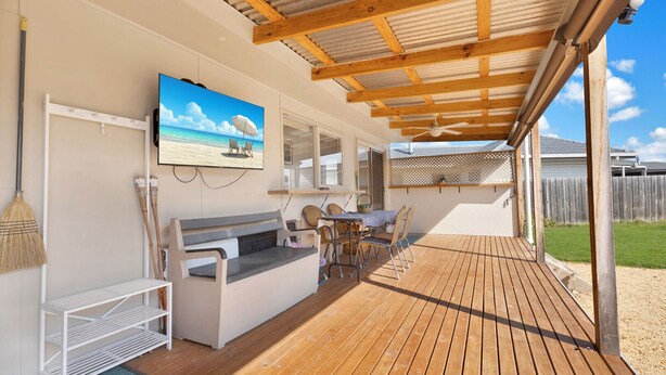A covered outdoor patio with a wooden deck, a seating area with a table and chairs, a TV mounted on the wall, and a view of the beach through a window.