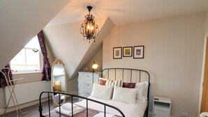 A bedroom with a sloped ceiling, a bed with white and brown bedding, a bedside table, a nightstand, a dresser, a mirror, and a chandelier.