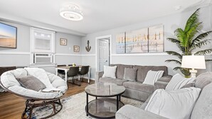 Een woonkamer met een grijze bank, een ronde salontafel en een rieten fauteuil. Er staat een eettafel met stoelen, een grote plant en kunst aan de muur.
