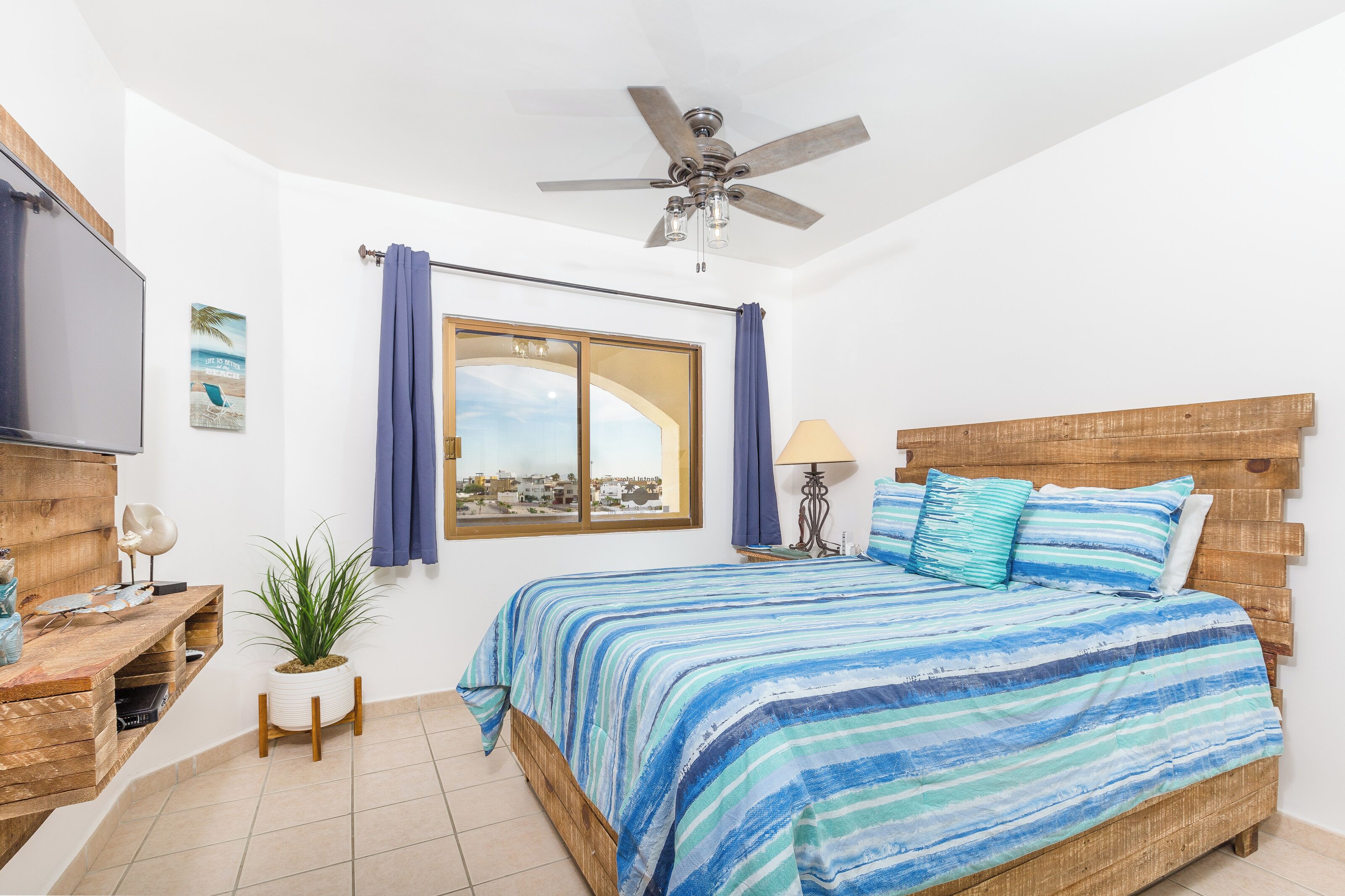 Une chambre à coucher avec un lit, un ventilateur de plafond, une fenêtre avec des rideaux, une table de chevet, une lampe et une plante en pot.
