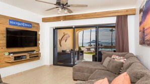 A living room with a flat-screen TV mounted on a wooden wall, a ceiling fan, and a large window with a view of the beach. - BeachBumCondos (Puerto Peñasco)