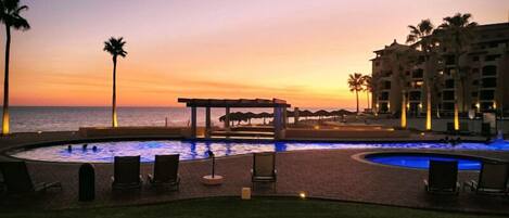 Un espace piscine avec des chaises longues, un coin salon abrité et une vue sur l’océan au coucher du soleil.