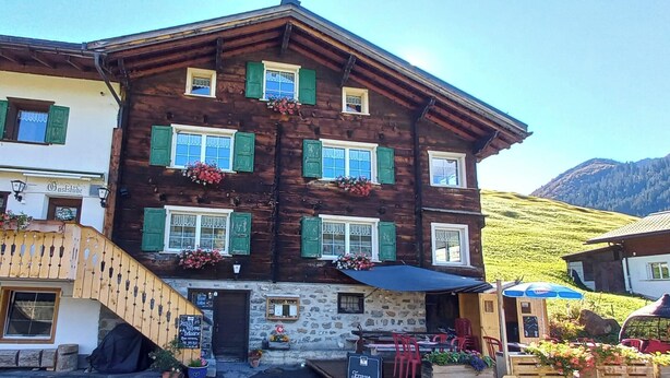 Un bâtiment traditionnel en bois, doté de nombreuses fenêtres, d’une façade en pierre et d’un espace extérieur aménagé avec des chaises rouges.