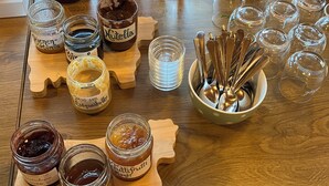 Une table en bois sur laquelle sont disposés divers aliments et condiments, notamment des pots de Nutella, du miel et des confitures, ainsi qu’une boîte de café et un verre de jus d’orange.