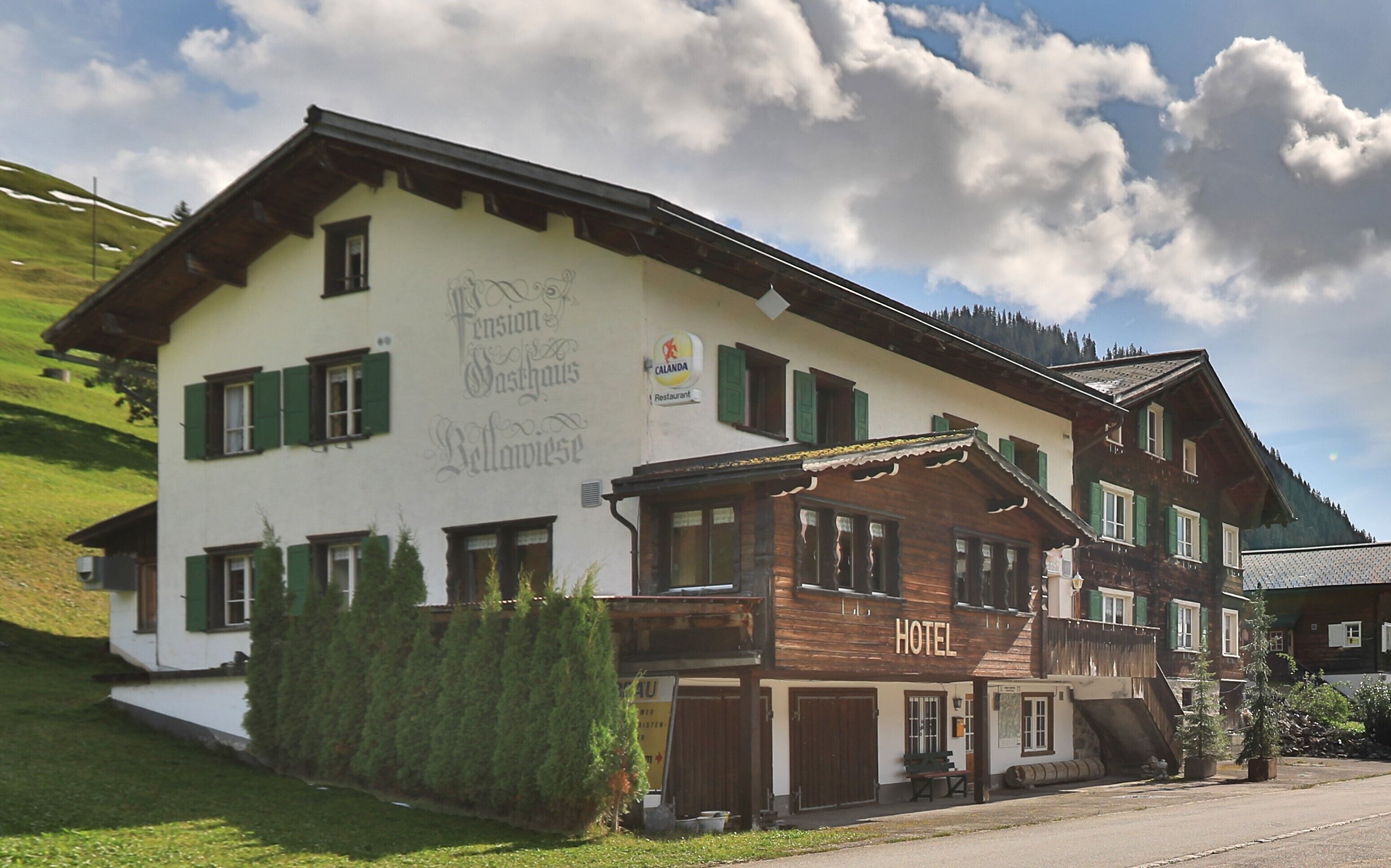 A traditional Alpine hotel with a white facade and green shutters, a visible restaurant sign, and a wooden annex labeled "HOTEL".