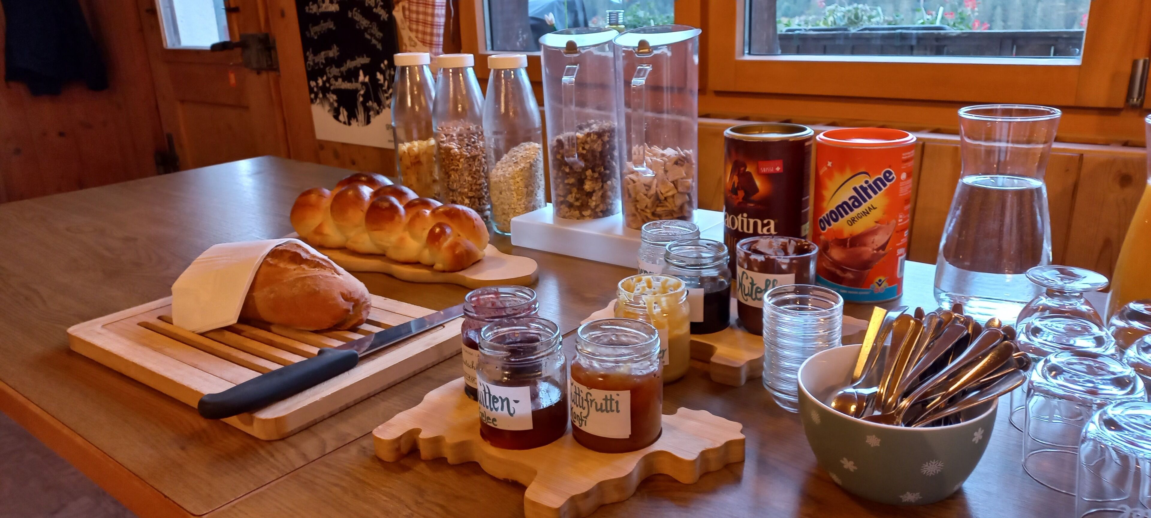 A wooden table with a loaf of bread, jars of preserves, a can of Evomarine, and various utensils.