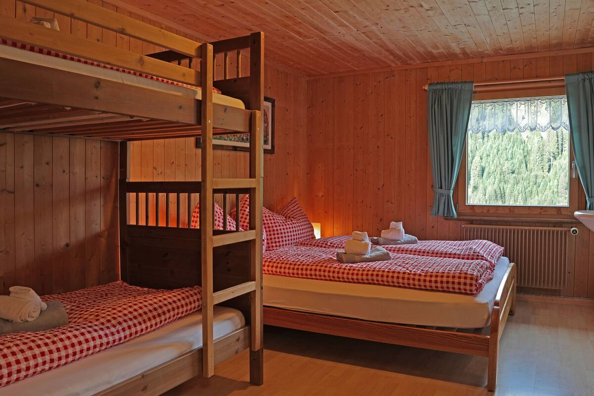 Uma cabana de madeira com beliches e uma cama simples, uma janela com cortinas e um radiador.