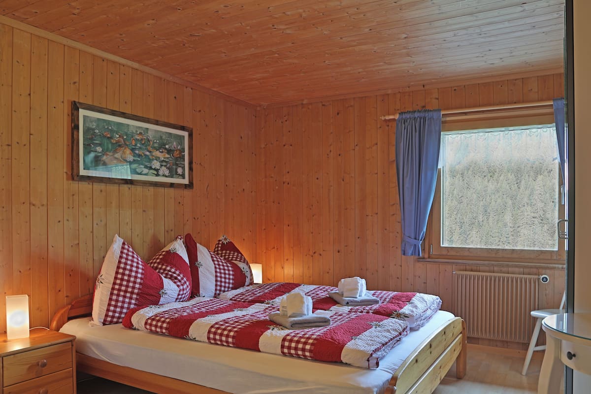 Quarto com painéis de madeira, uma cama, almofadas, uma mesa de cabeceira, uma janela com cortinas e um quadro na parede.