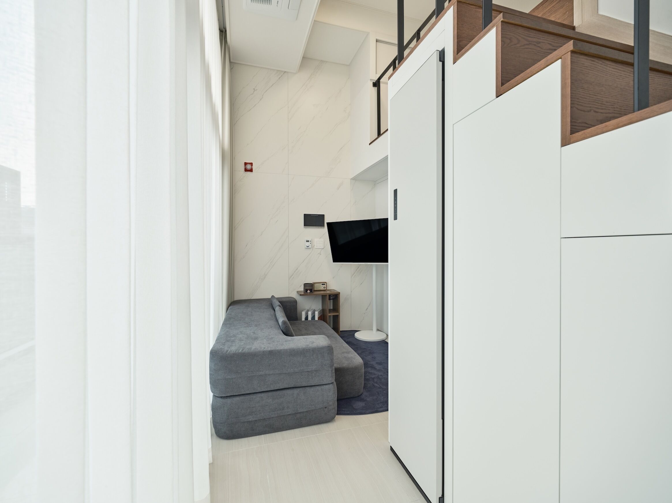 A modern, compact room with a grey sectional sofa, a flat-screen TV mounted on the wall, and a small side table with a lamp.