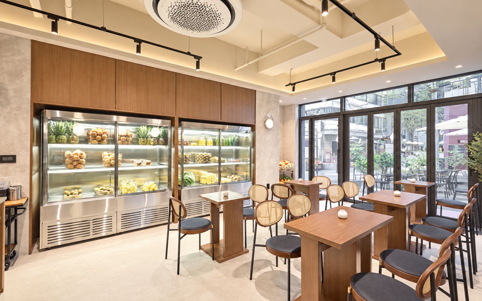Interior de una cafetería moderna con mobiliario de madera, vitrinas de vidrio con alimentos y grandes ventanales con vista a la calle.