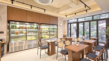 Interior de una cafetería moderna con mobiliario de madera, vitrinas de vidrio con alimentos y grandes ventanales con vista a la calle.
