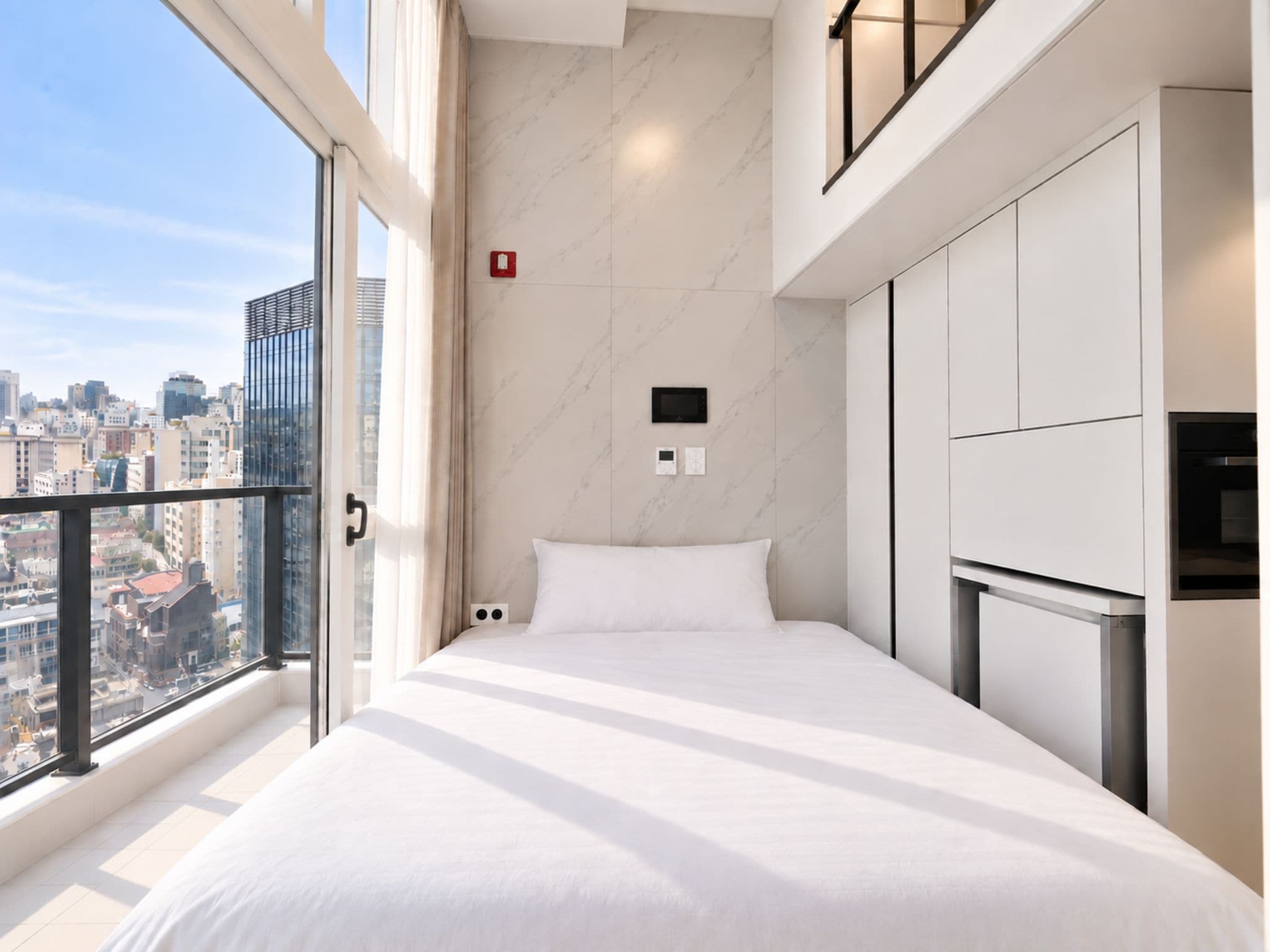 A modern bedroom with a city view, a bed, a built-in TV, and a small kitchenette.