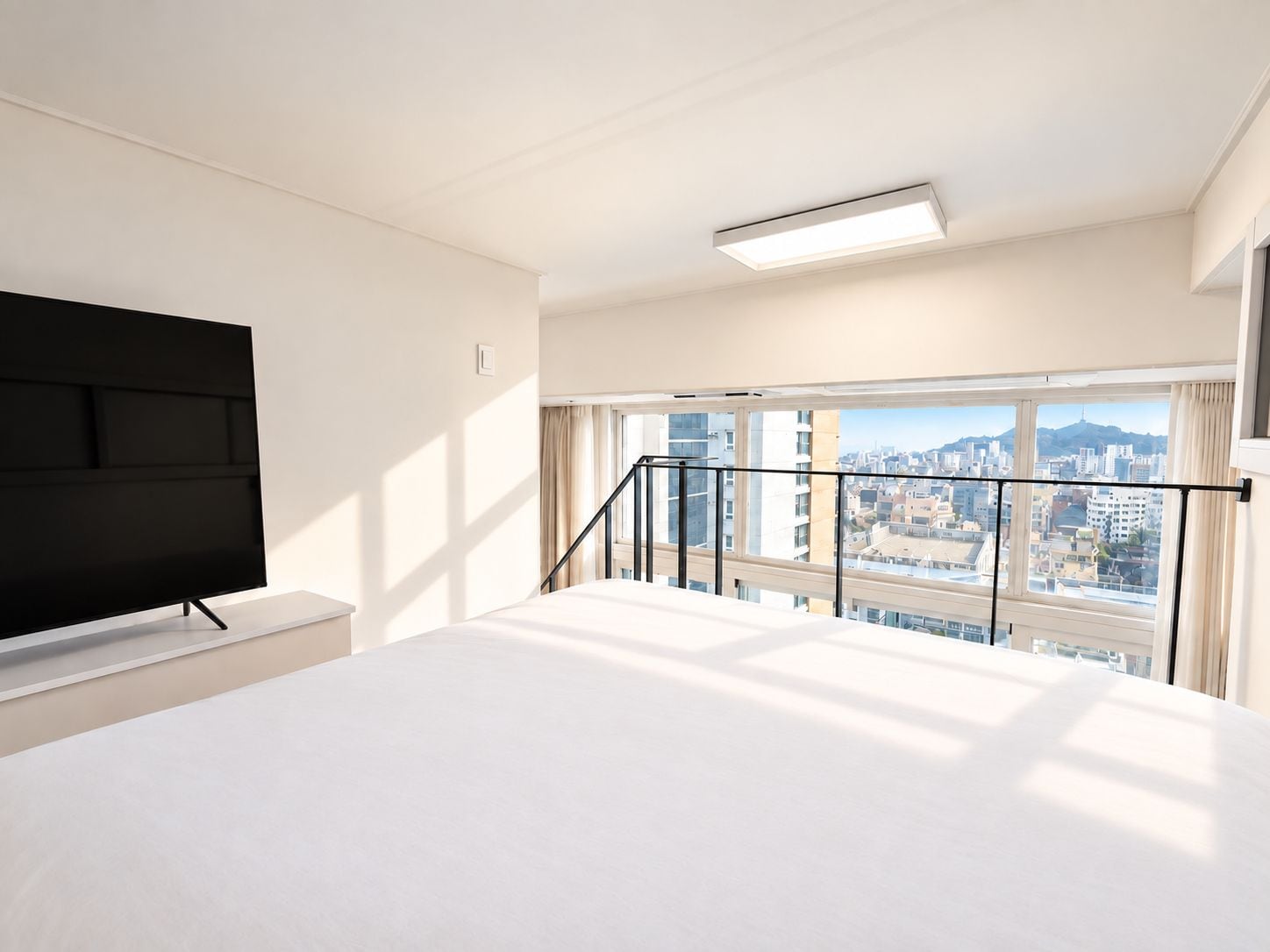 A hotel room with a bed, a flat-screen TV, a window with a city view, and a balcony railing.