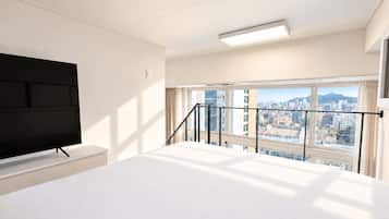 A hotel room with a bed, a flat-screen TV, a window with a city view, and a balcony railing.