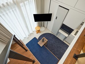 Habitación de hotel moderna con una televisión de pantalla plana montada en la pared, una mesa redonda pequeña, una alfombra azul y un taburete de madera.