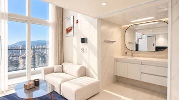 A modern hotel room with a sofa, a small table with a radio, a large window with a city view, a bathroom with a mirror, and a marble floor.