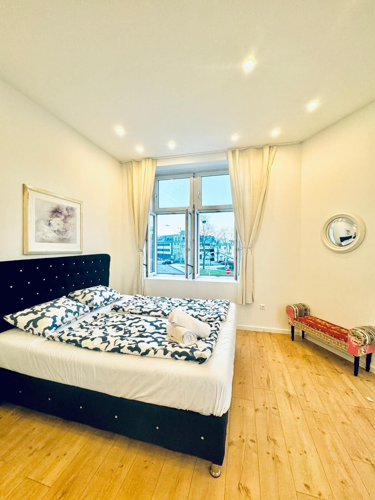 Une chambre à coucher avec un lit, une fenêtre avec des rideaux, une table de chevet et un miroir au mur.