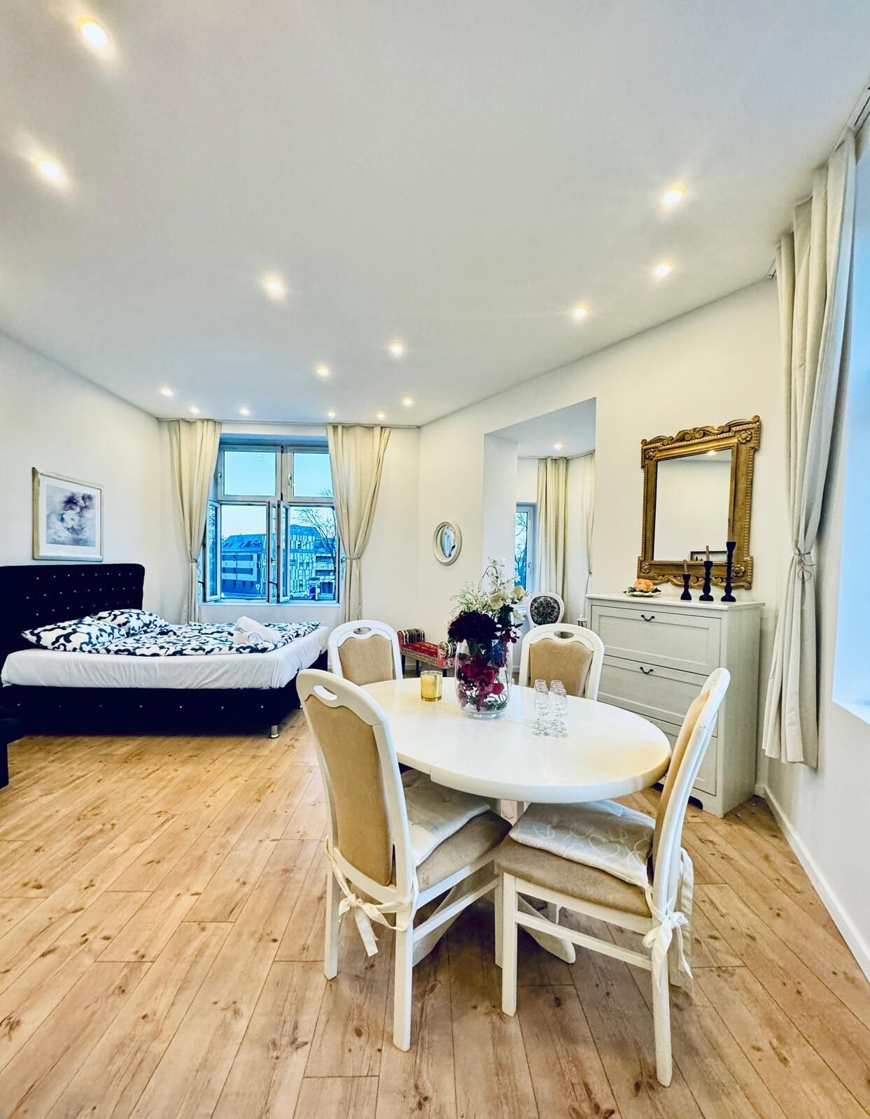 A modern bedroom with a bed, a dining table with chairs, a dresser, and a large mirror.