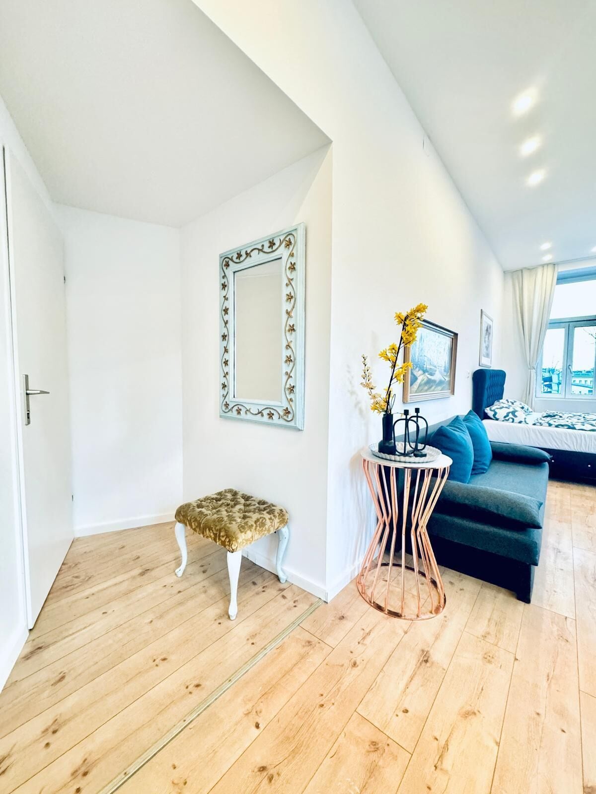 A modern living room with a wooden floor, a blue sofa, a decorative stool, and a round table with a vase of flowers.