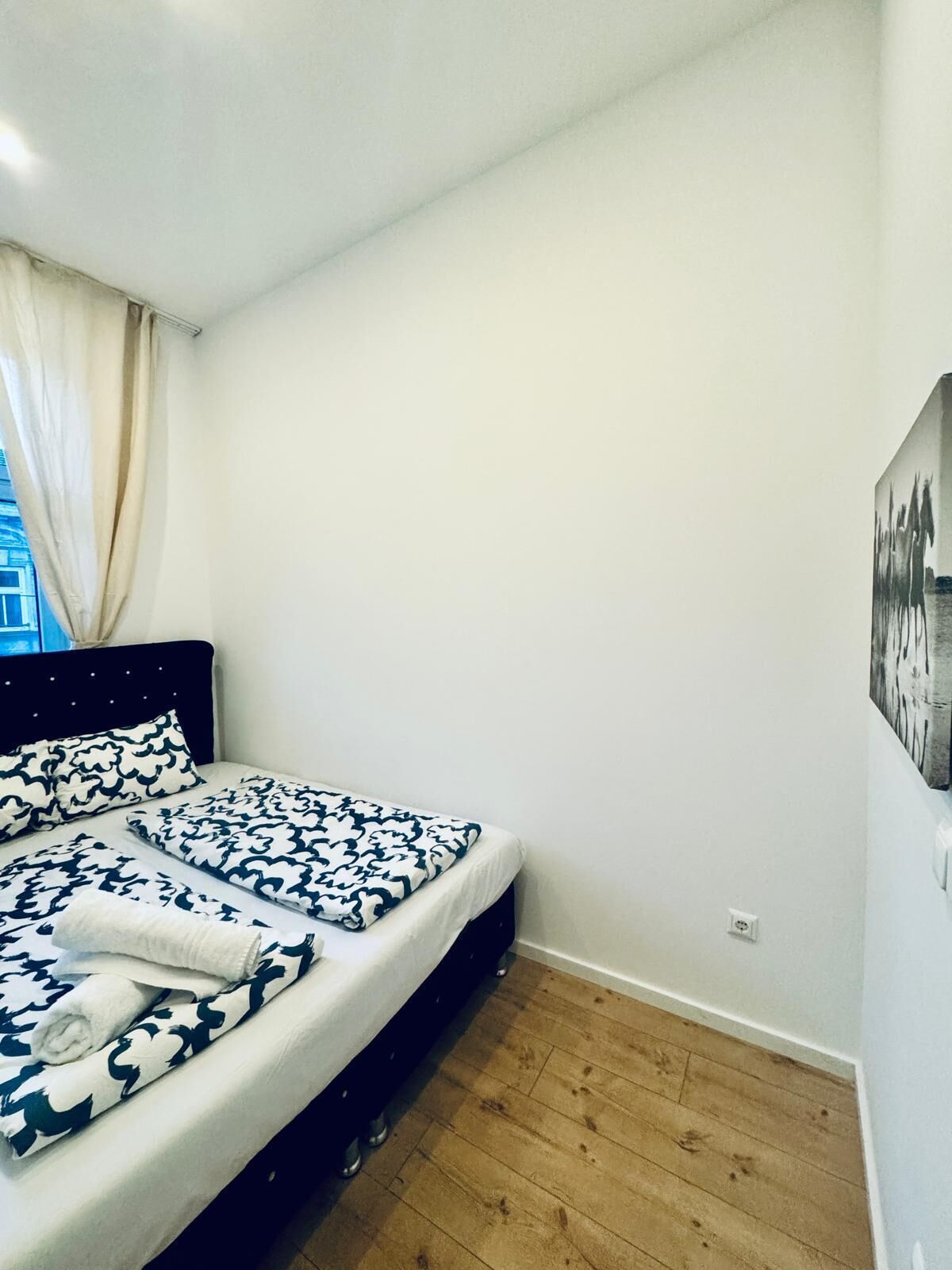 A bedroom with a bed, a headboard, a window with curtains, and a wall-mounted artwork.