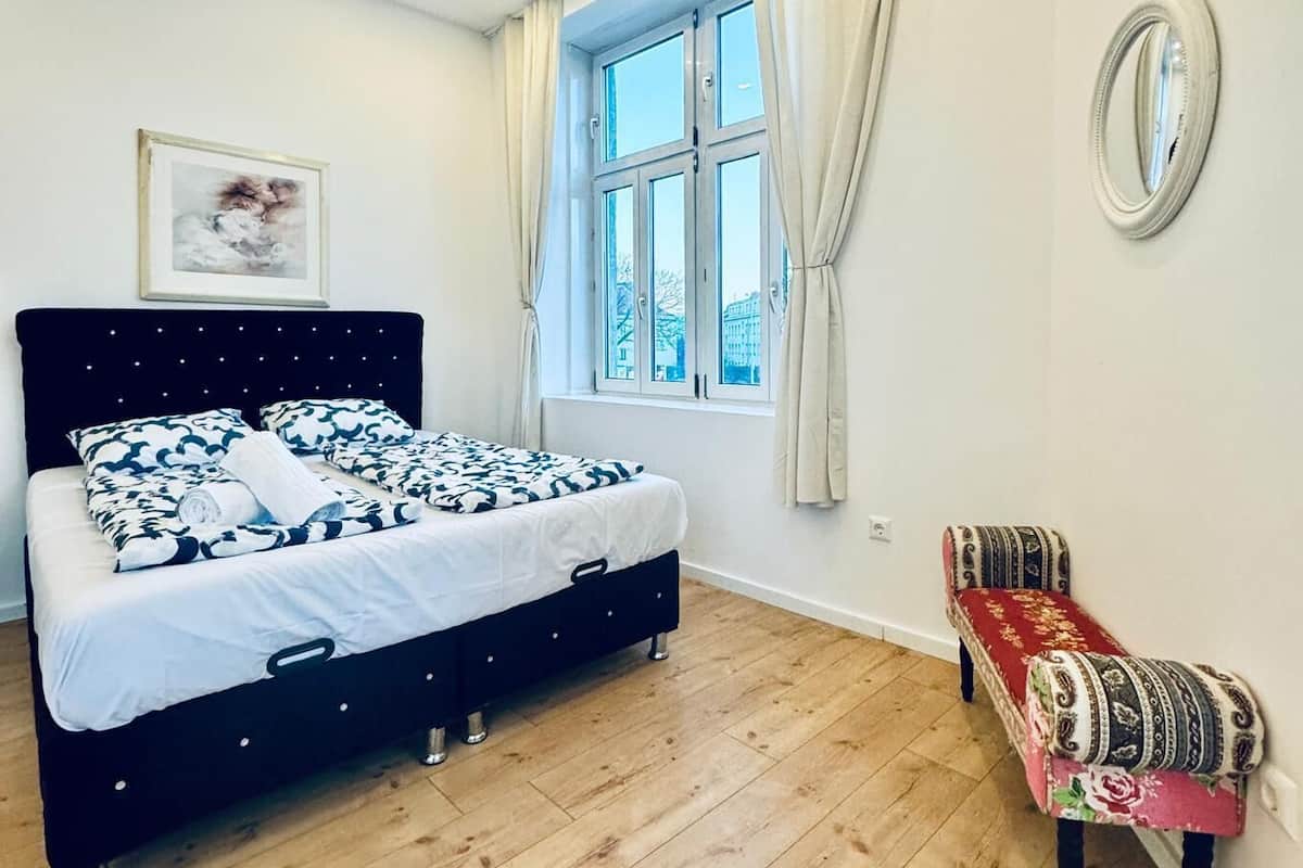 A bedroom with a bed, a window with curtains, a small bench, and a wall-mounted mirror.