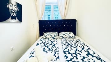 Un dormitorio con cama, cortinas, un cuadro y ventana con vistas a edificios.